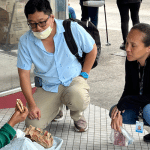 Two people consult a person sitting on the ground eating a sandwich.